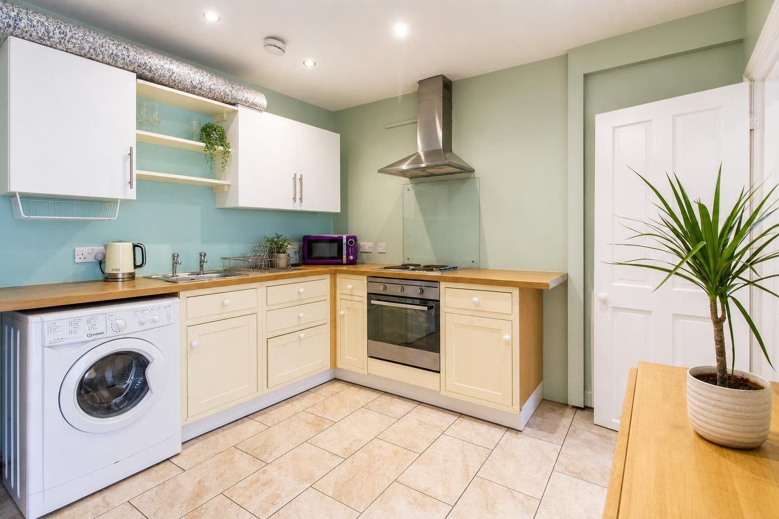 Bright fitted kitchen with cream cabinets, wooden countertops, a washing machine, oven, hob and potted plant.