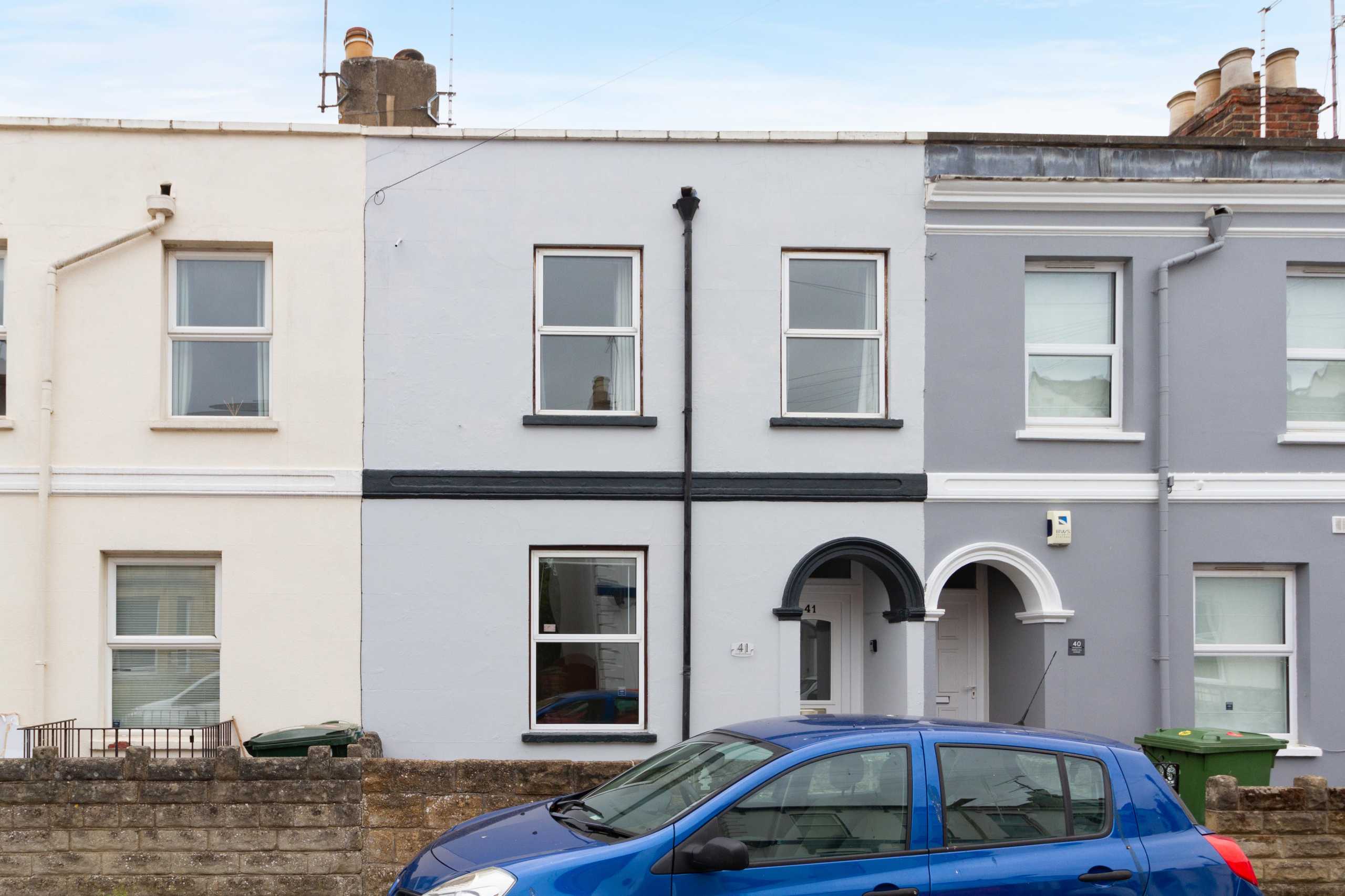 Street view of adjoining terraced houses with painted façades, windows, arched entrances and a blue car in front.