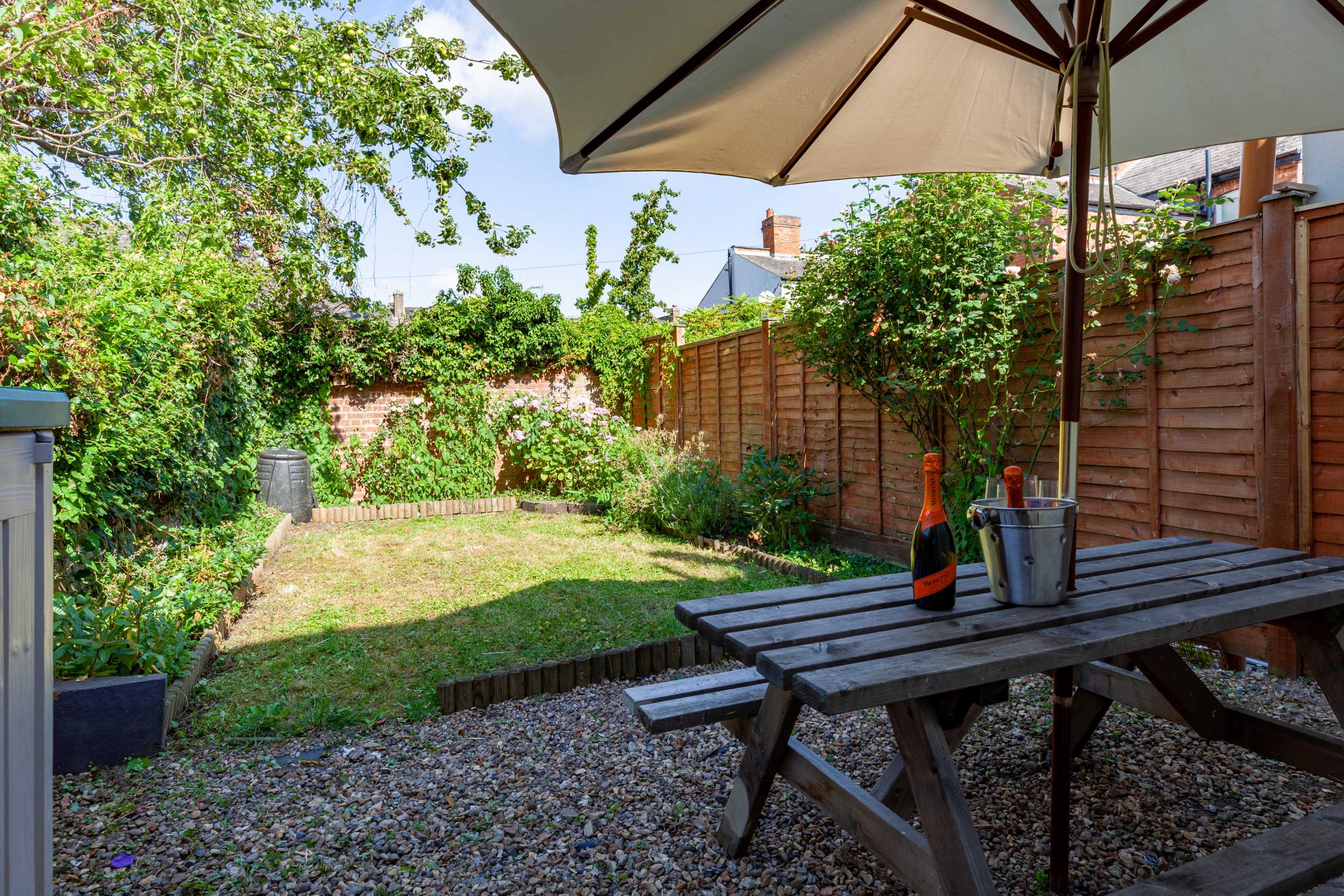 A fenced garden with grass, plants, gravel, and a wooden picnic table beneath a large umbrella.