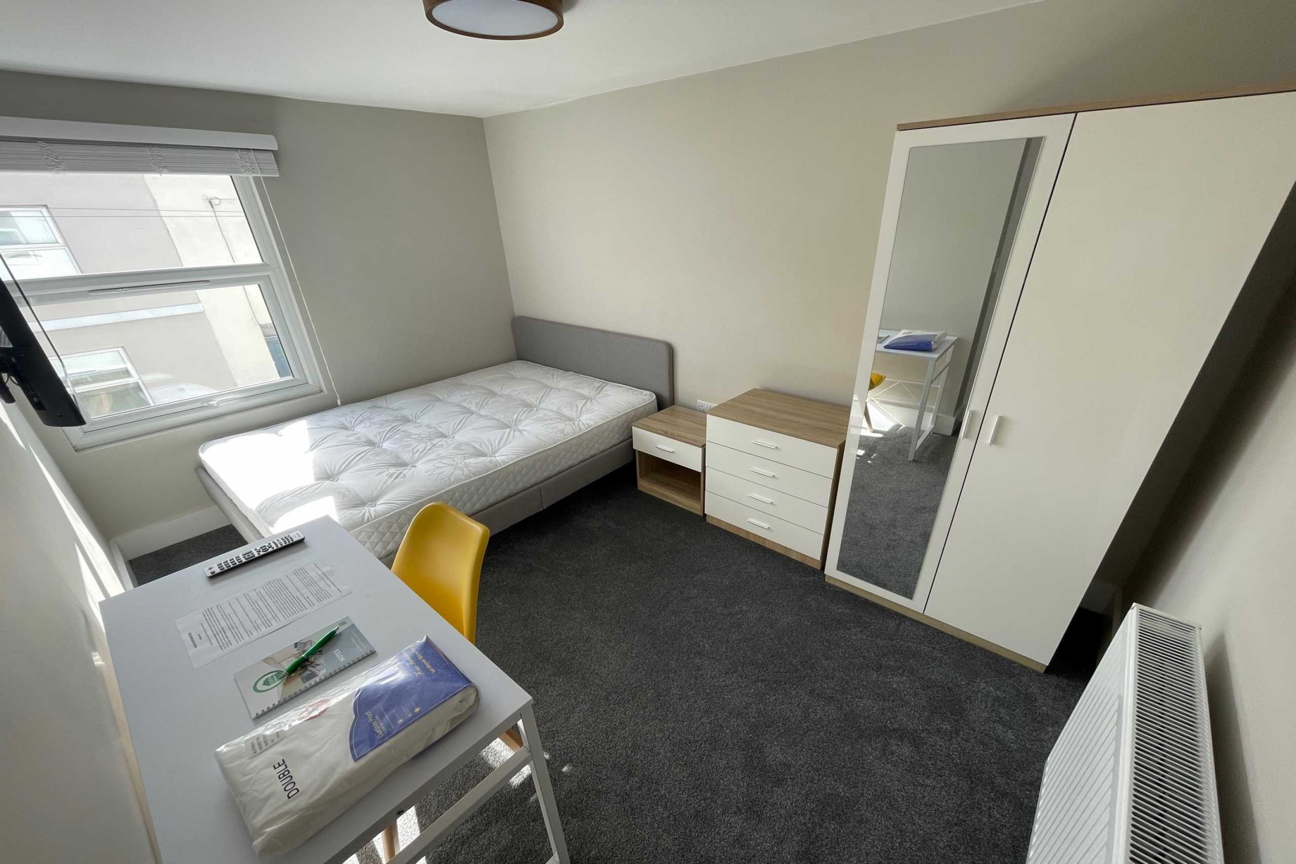 A furnished bedroom with a single bed, desk, chair, chest of drawers, wardrobe and window.