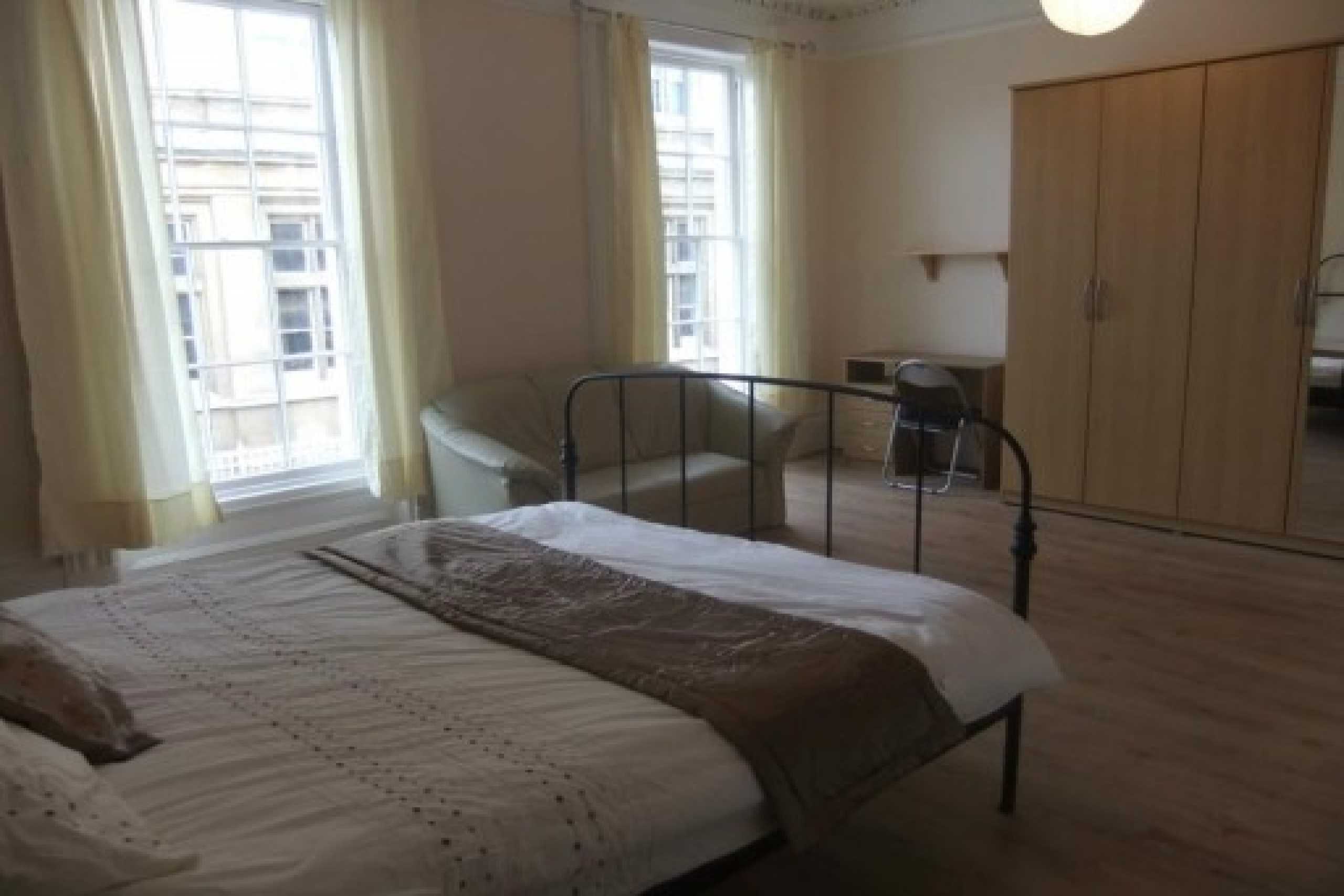 A bedroom with a double bed, tall windows, a wardrobe, an armchair and a small desk.