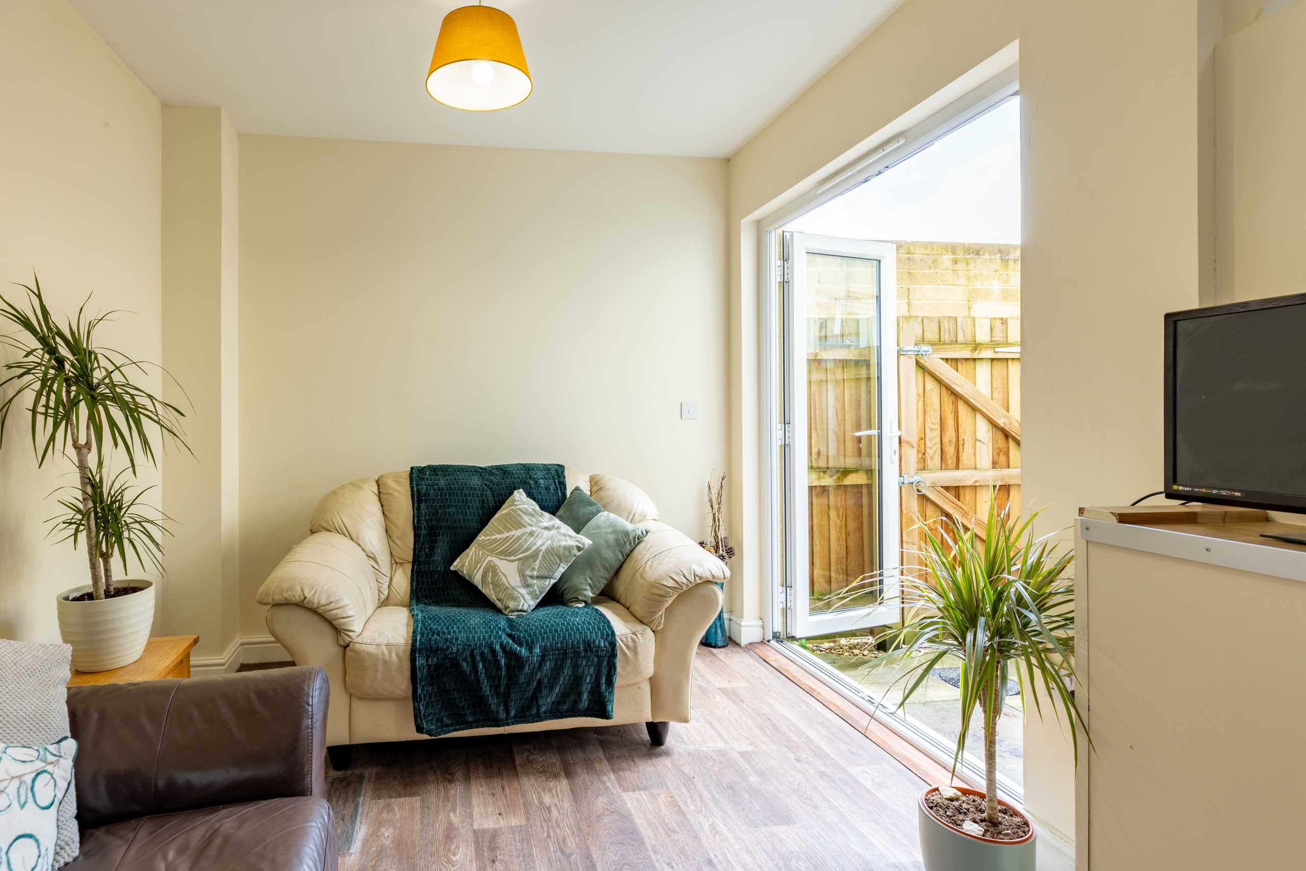 Bright sitting area with an armchair, sofa, television, potted plants and an open glass door to a fenced outdoor area.
