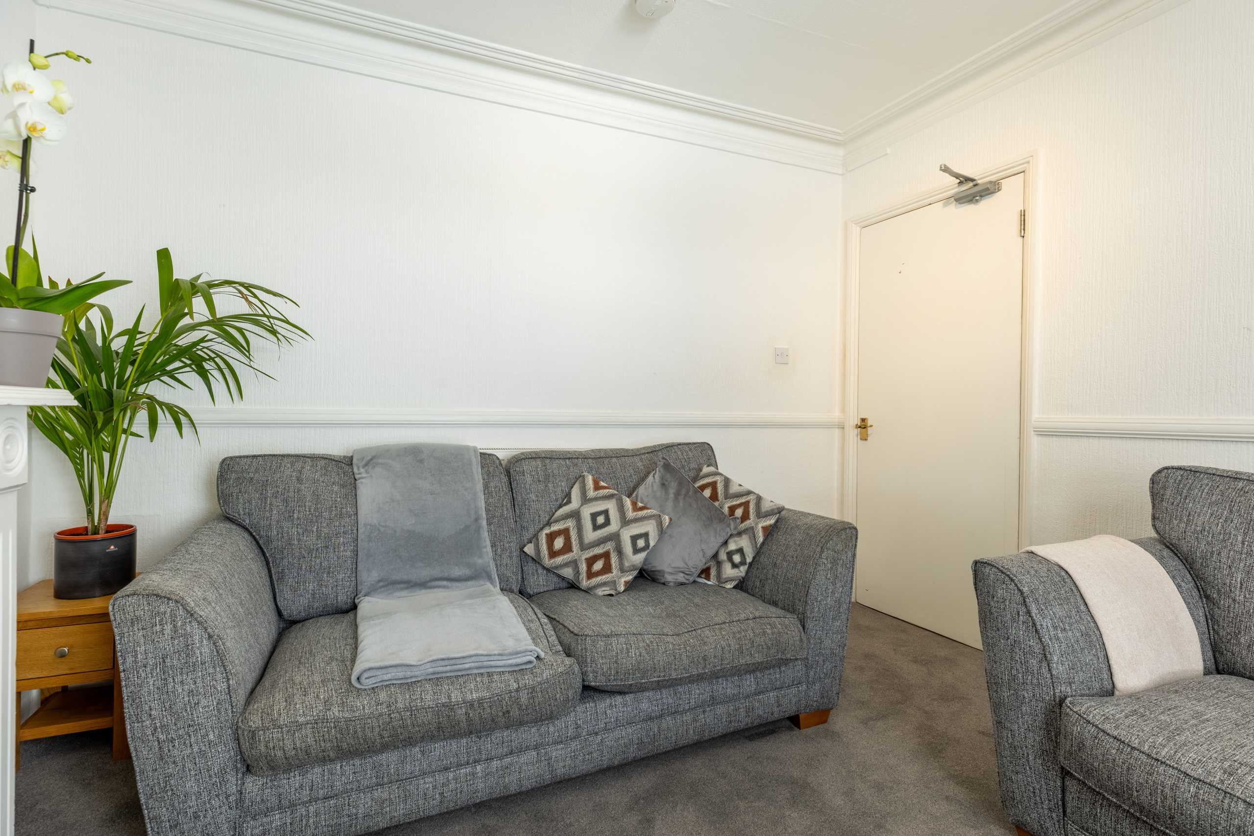A furnished living room with grey sofas, patterned cushions, a small side table, a potted plant and a closed door.