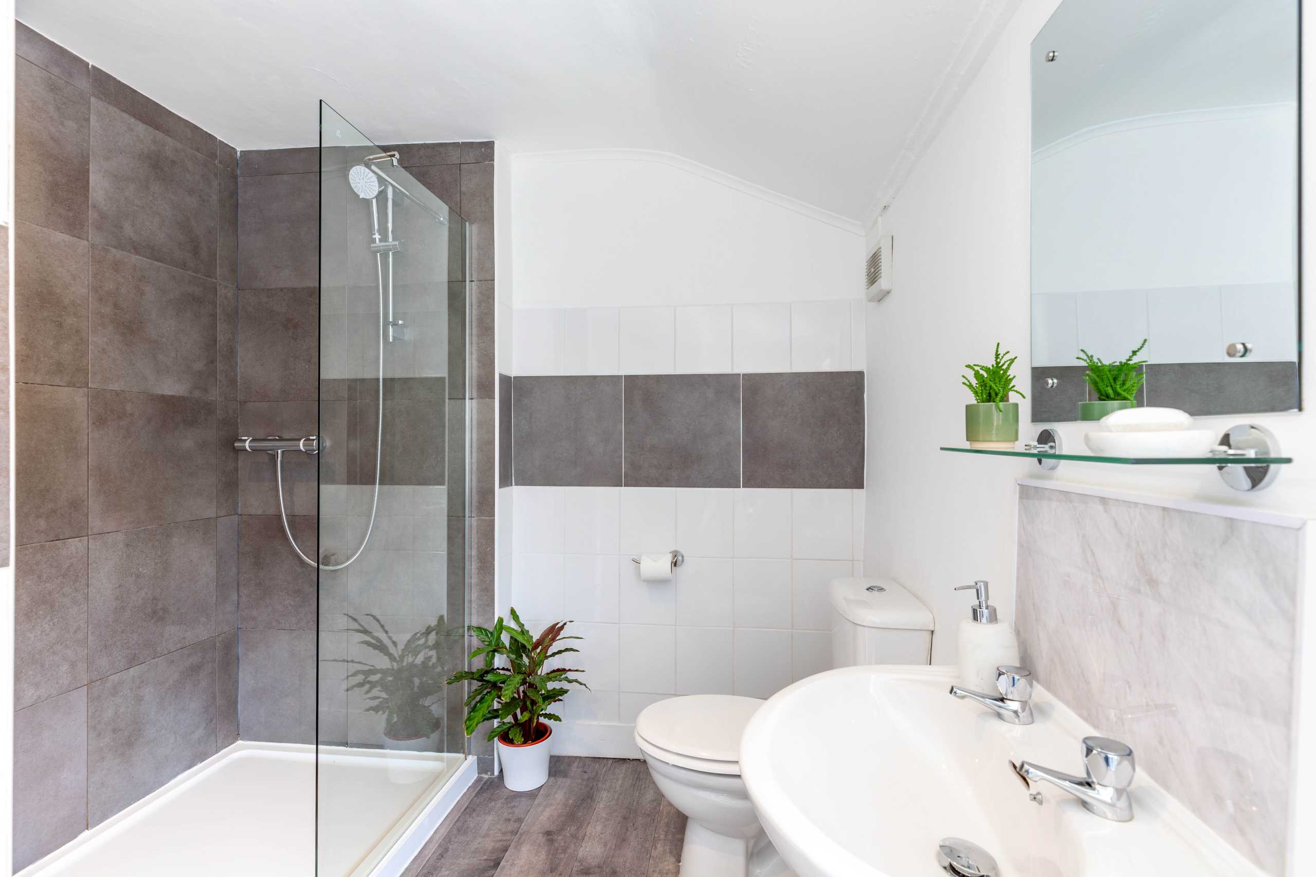 Bright modern bathroom with a glass shower enclosure, toilet, pedestal sink, tiled walls and potted plants.