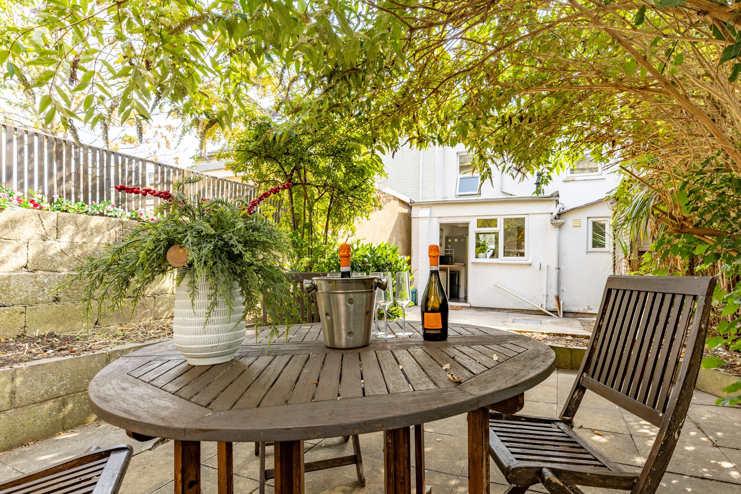 Garden patio with a round wooden table, chairs, plants and a white house in the background.