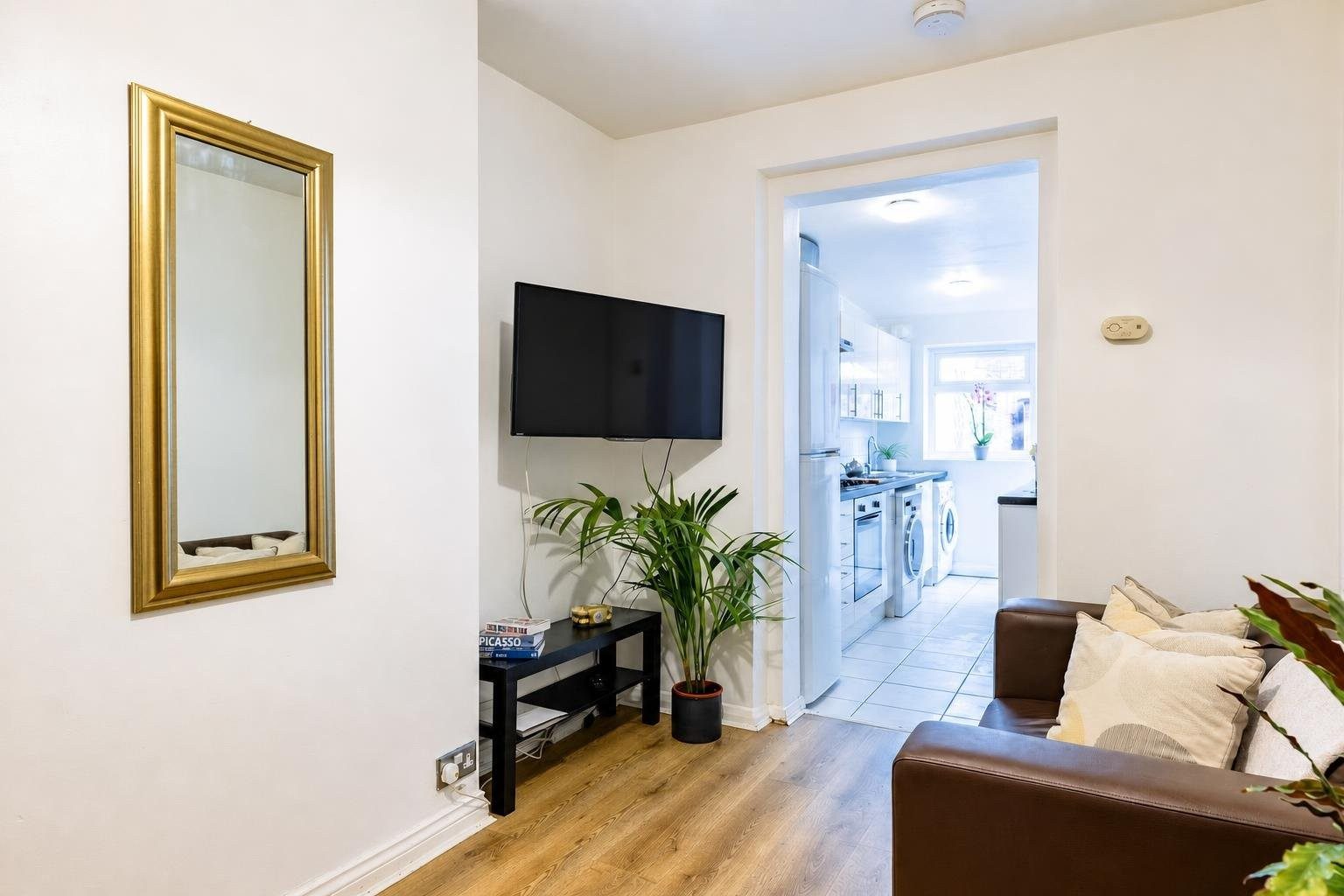 Living room with a wall-mounted television, sofa, mirror and doorway leading to a bright kitchen.