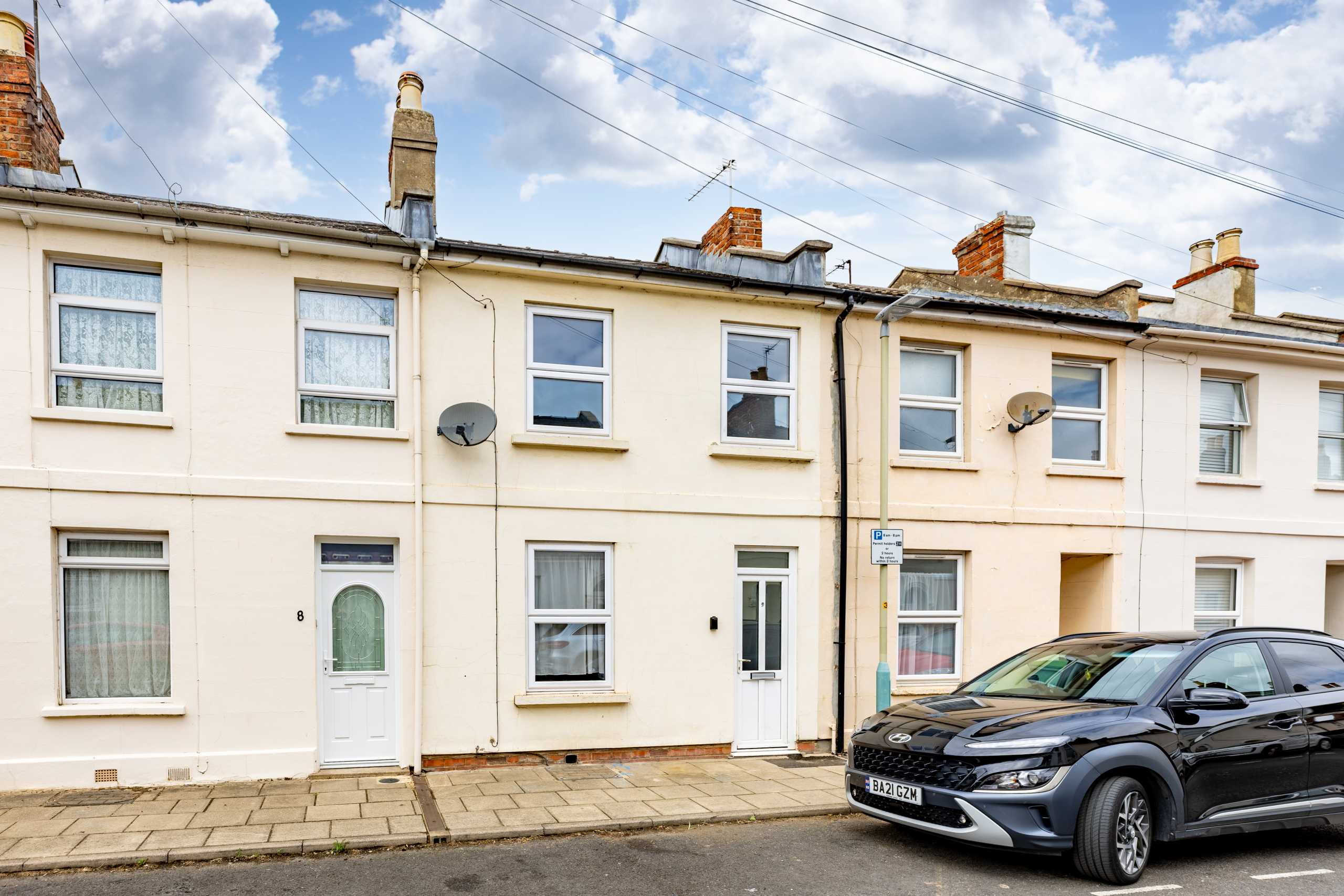 Row of attached terraced houses with front doors, windows, chimneys and parked cars along the street.